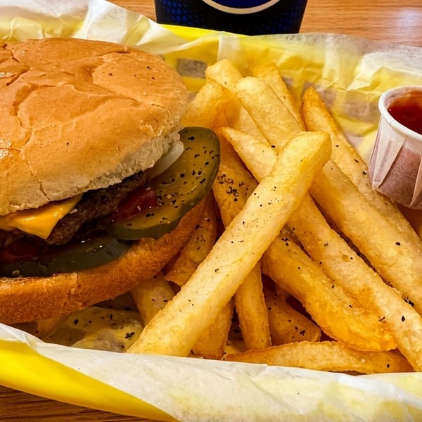 Cheeseburger with pickles and fries in a yellow basket, with two ketchup cups.