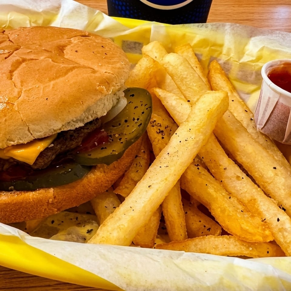 Cheeseburger with pickles and fries in a yellow basket, with two ketchup cups.