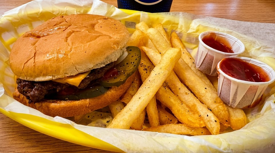 Cheeseburger with pickles and fries in a yellow basket, with two ketchup cups.